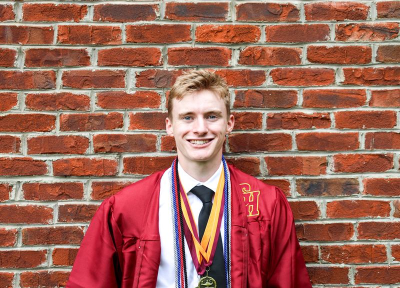 Graduate in a red cap and gown against a brick wall